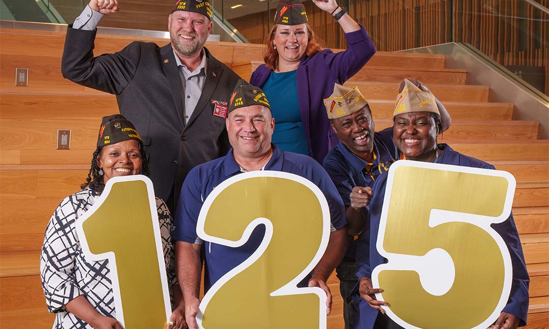 VFW members with raised hands holding a sign for the 125th anniversary of the organization